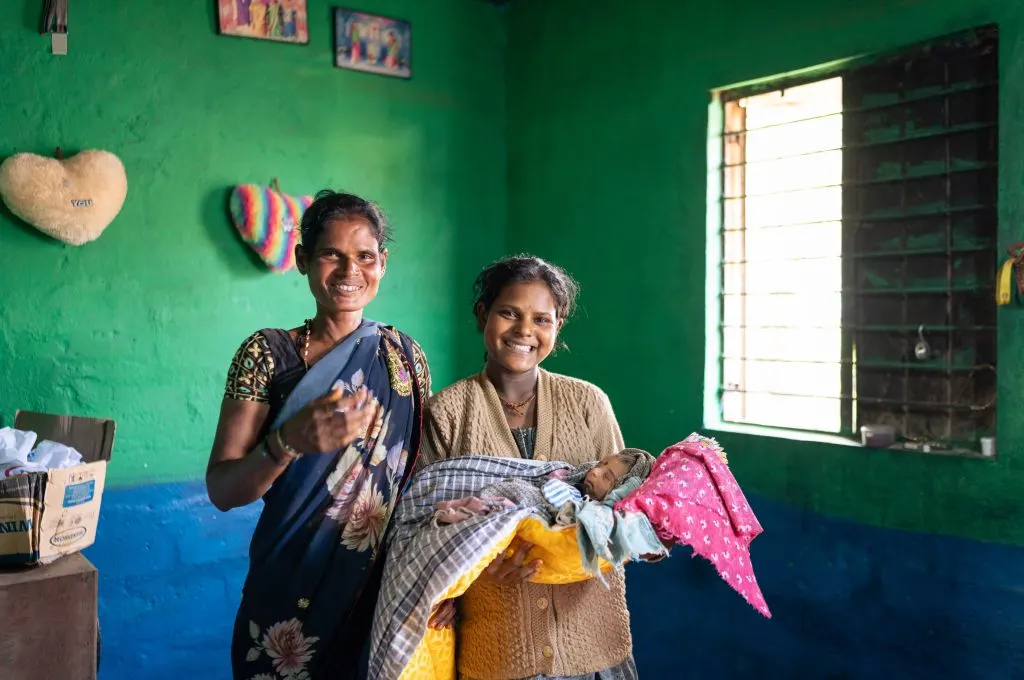 The image features a woman posing with a young girl, presumably a family member. The girl is holding a baby in her arms. The woman and the young girl are smiling._Maternal and newborn care