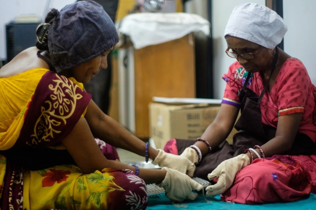 The image features two women sitting together and placing what appear to be small clear bottles on a small rack while wearing gloves._ONDC platform