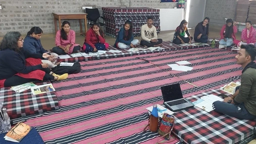 The image features a group of young people sitting on mattresses in a semi-circle. They are facing an older person who is addressing the group._Social norms