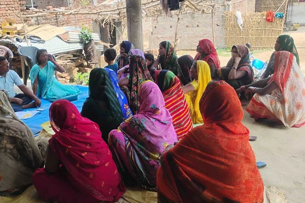 A group of rural women, dressed in colorful traditional sarees, sit on the ground in a semi-circle, attentively listening to a woman in a blue saree speaking during a community meeting held outdoors in a village_microfinance India