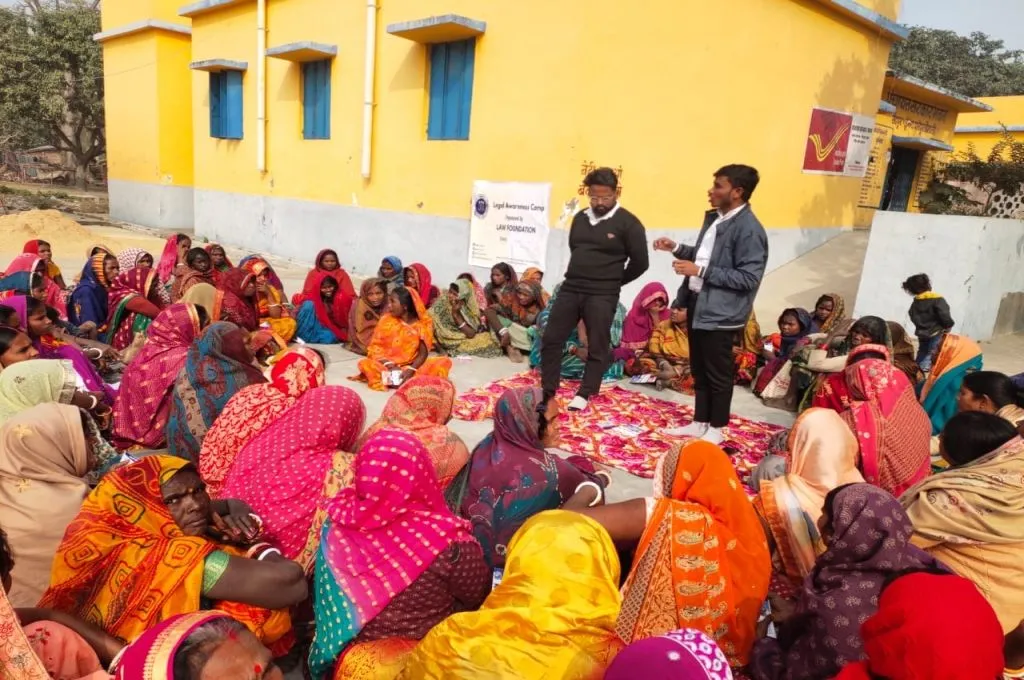 The image is set in a rural area and features a group of women sitting on the ground in a circle outside a building. Two young men stand in the middle of the circle and address the gathering. The wall of the building behind the group has a banner stuck to it which reads 'Legal Awareness Camp organised by Law Foundation'._Criminal justice