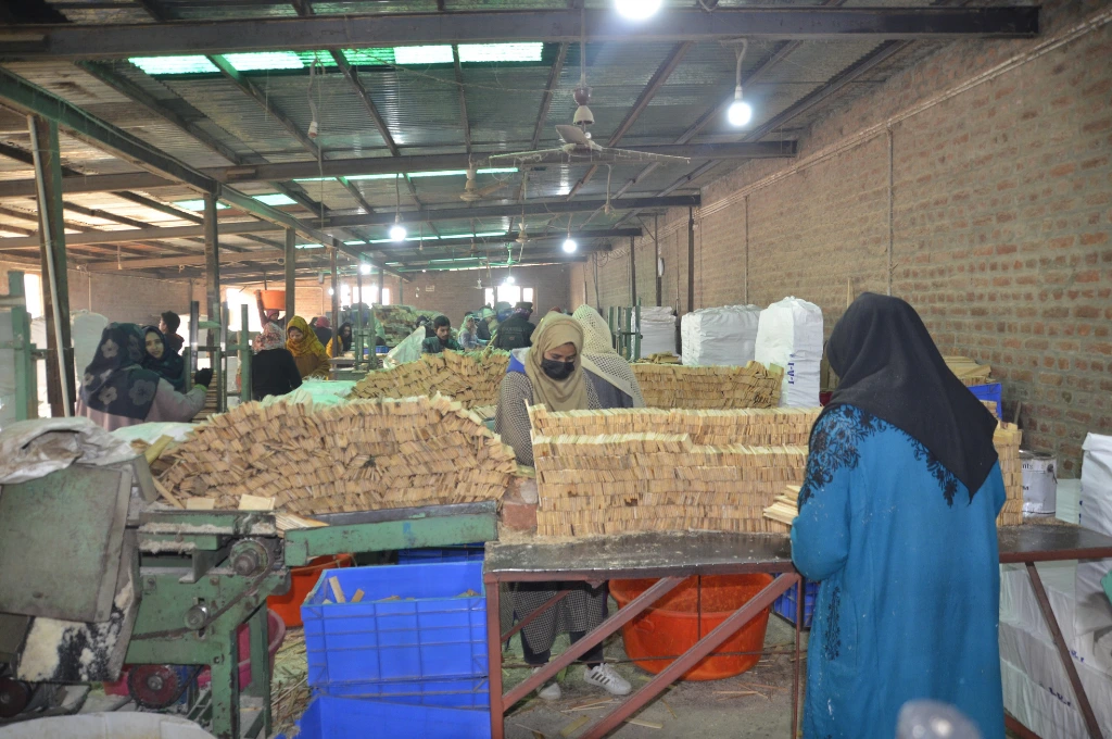 The image features the inside of a pencil-manufacturing facility with brick walls and a tin roof. The workers are mostly women. In the foreground, two women are stacking small slats of wood into rows on a metal table. A machine and plastic crates are placed on the floor._Oukhoo pencil village 