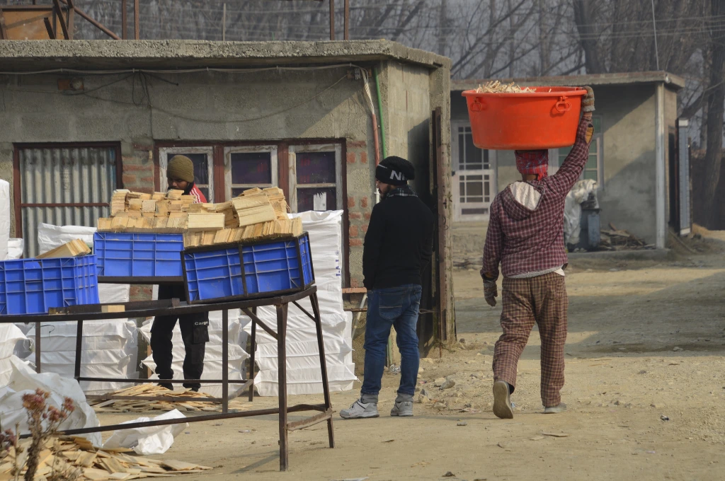 The image shows a metal table with plastic crates placed on it out in the open. Each crate contains small wooden slats. A man is seen placing another crate on the table while another man stands next to him. A third man walks past them carrying a plastic tub of what appears to be fodder on his head. There are small room-like concrete structures in the background._Oukhoo pencil village 
