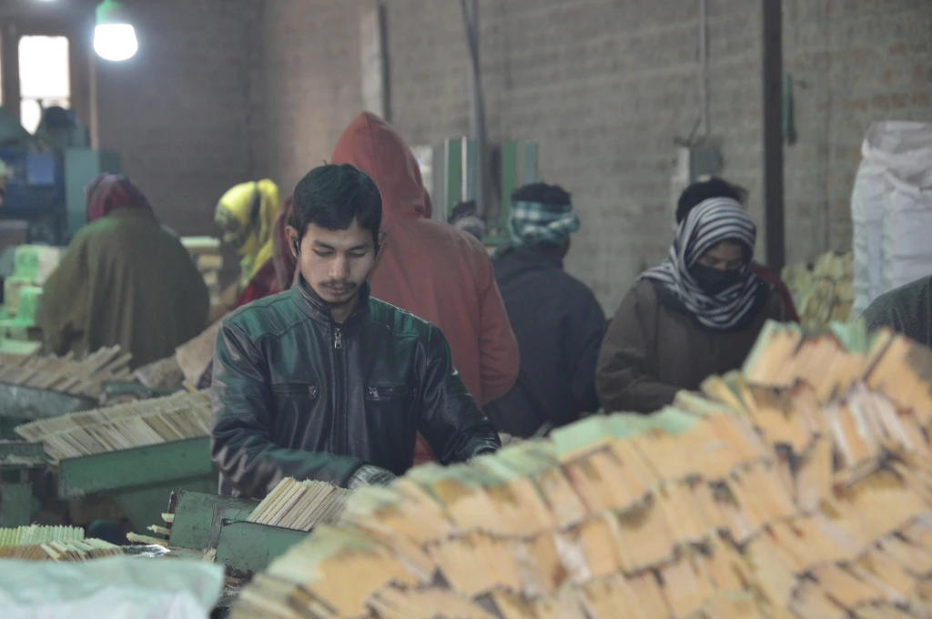 This is another image of a pencil-manufacturing facility. A young man sets slats of wood in a metal machine. Behind him, other workers, including women, similarly arrange wooden slats._Oukhoo pencil village 
