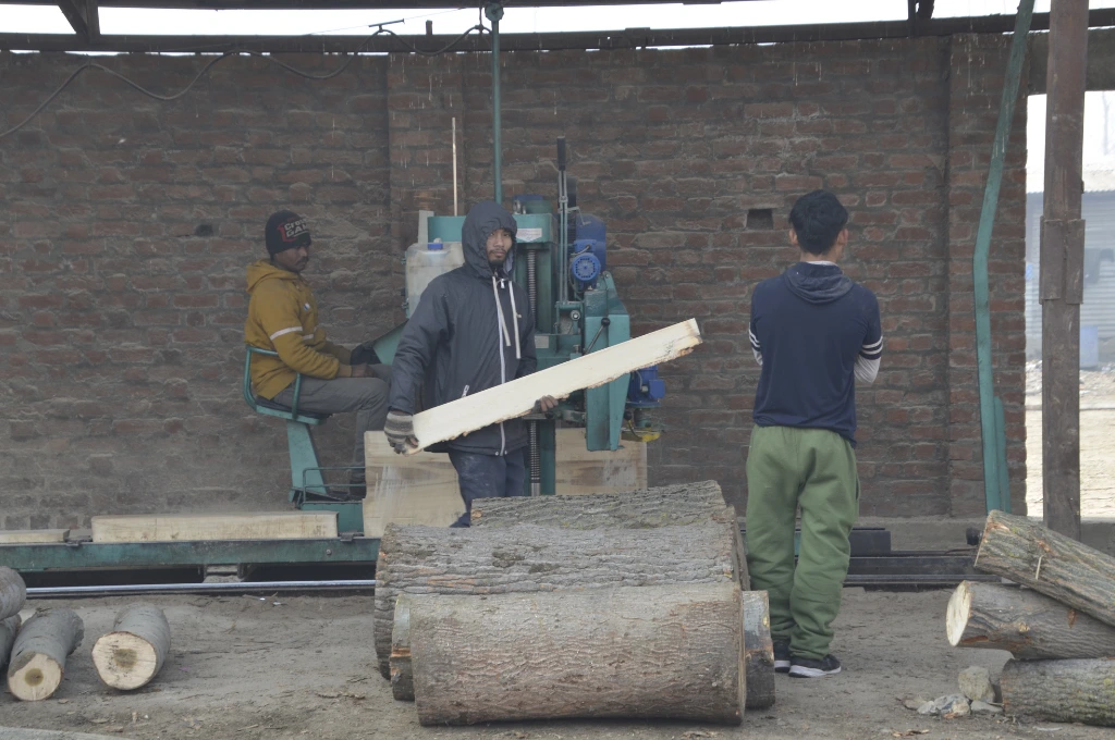 The image depicts a group of three men. One man is seated and operating a big machine. The second man is holding a rectangular piece of wood in his hands, while the third man stands with his back turned. There are big logs of chopped wood placed on the ground._Oukhoo pencil village 