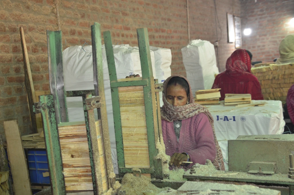 The image is of the inside of a pencil-manufacturing facility. It depicts a women seated in front of a table which has a metal frame with slats of wood stacked in it. There is a second metal frame with wooden slats behind it. The table is covered in piles of sawdust. There is another woman standing in the background with her back turned, and there are big white bags with packaged materials placed next to the brick wall._Oukhoo pencil village 