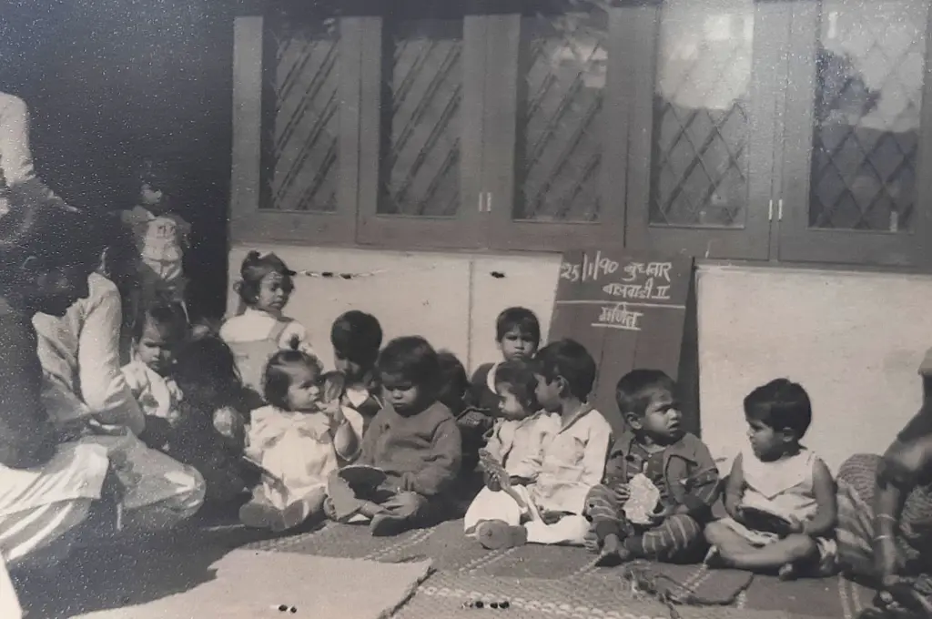 The image is a black and white photo of a group of young children sitting on a rug on the ground outside a room. A blackboard leans against the wall behind the children. A young woman is sitting with the children._Early childhood care
