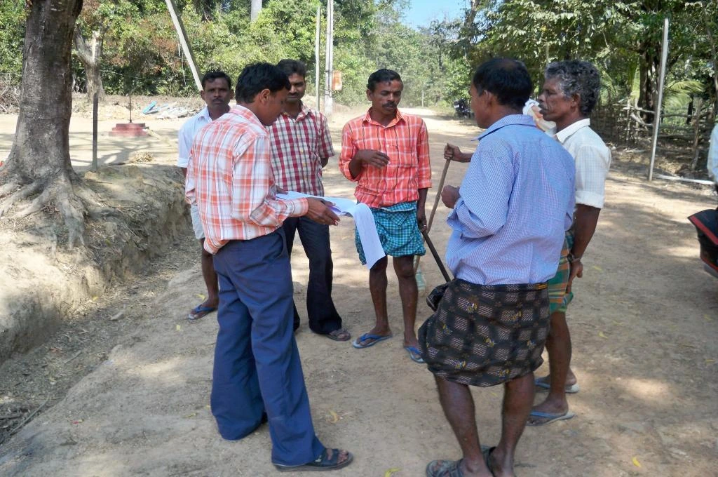 The image features a group of men standing in a circle on an unpaved road and talking. One of the men is holding sheets of paper in his hand._data quality
