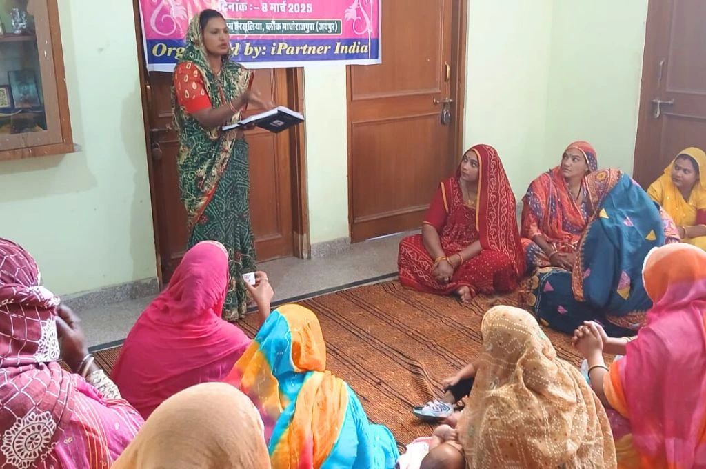 A group of women in colorful sarees are gathered in a small room for a community meeting or workshop. One woman, standing near a banner on the wall, is speaking and holding a notebook, while the others sit on a mat on the floor, listening attentively_Nat community