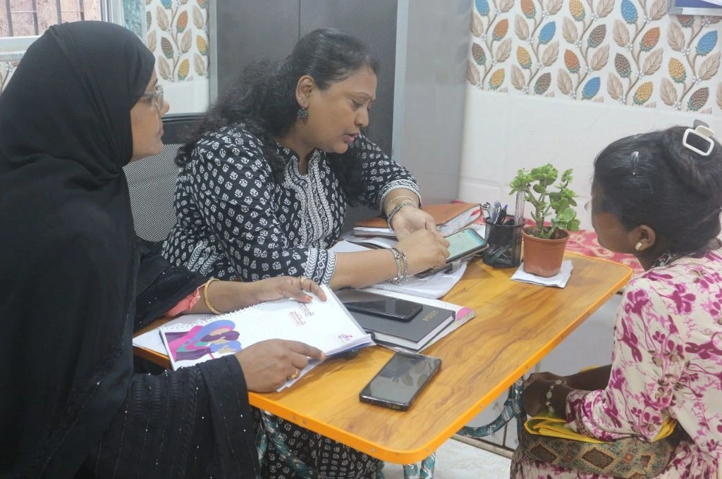 Three women are seated around a small wooden desk in an office setting. One woman in a black and white patterned kurta is showing something on a mobile phone to the other two. One woman, wearing a black hijab, holds a booklet, while the third woman, dressed in pink floral attire, listens attentively. The desk is cluttered with notebooks, a phone, and a small potted plant. The background features a wall with a colorful leaf-patterned wallpaper_domestic violence