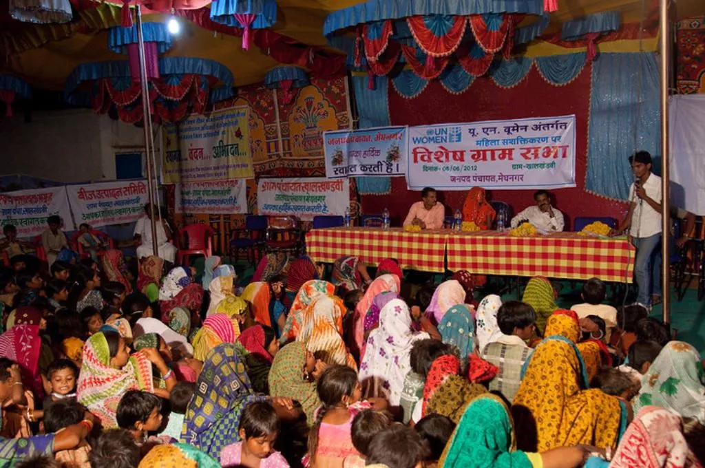 a gram sabha meeting with a panel sitting behind a table, and women in colourful chunnis sitting on the ground as participants-panchayati raj