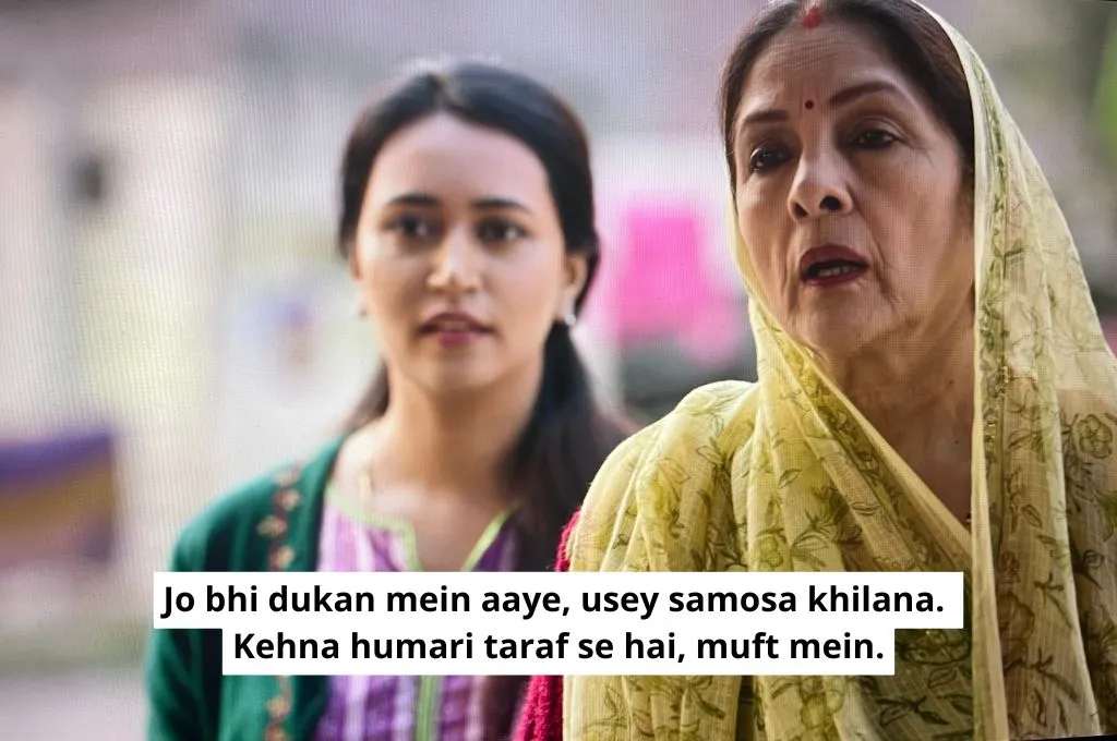 The image is a scene from Panchayat. An older woman, who is the village sarpanch, is telling someone, "Whoever comes to your shop, you give them a samosa to eat. And tell them that it is from us, for free". A younger woman, her daughter, stands in the background._Nonprofit humour