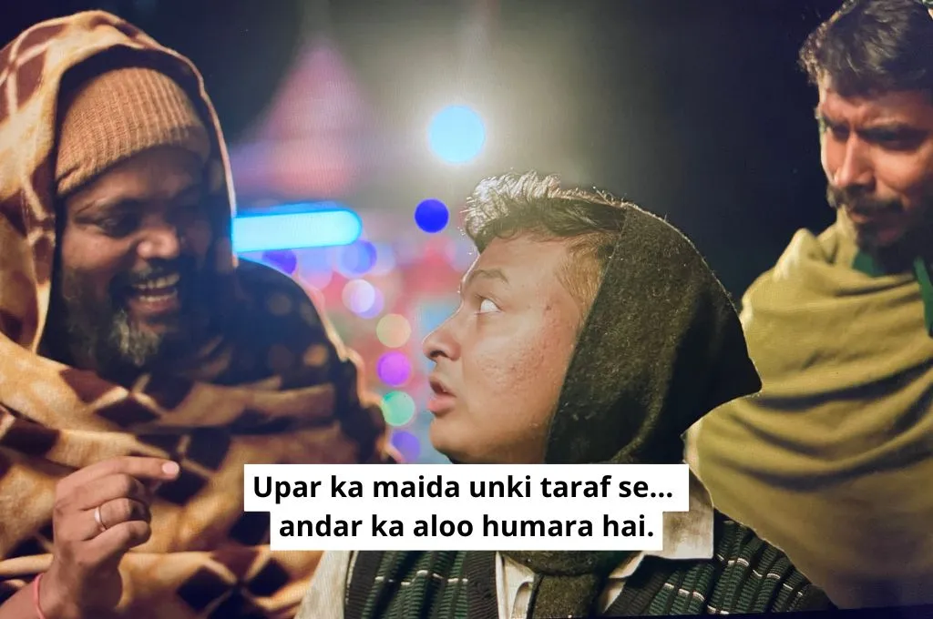 The image is a scene that is set in the same shop as the first image. This time, as a customer eats a samosa, two men who belong to a rival political group stand behind him. The older man tells the customer, "They (the sarpanch and her group) provided the flour to make the outer crust of the samosa, we provided the potatoes that went inside the samosa"._Nonprofit humour