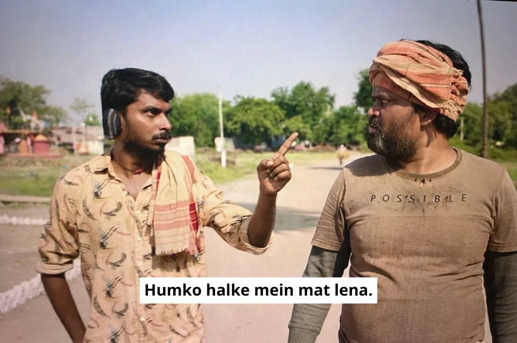 The image features the two men from the scene in the shop. They are the political rivals of the sarpanch's family. As both men walk down the road, the younger man points his finger at the older man and says in anger, "Don't take me lightly"._Nonprofit humour