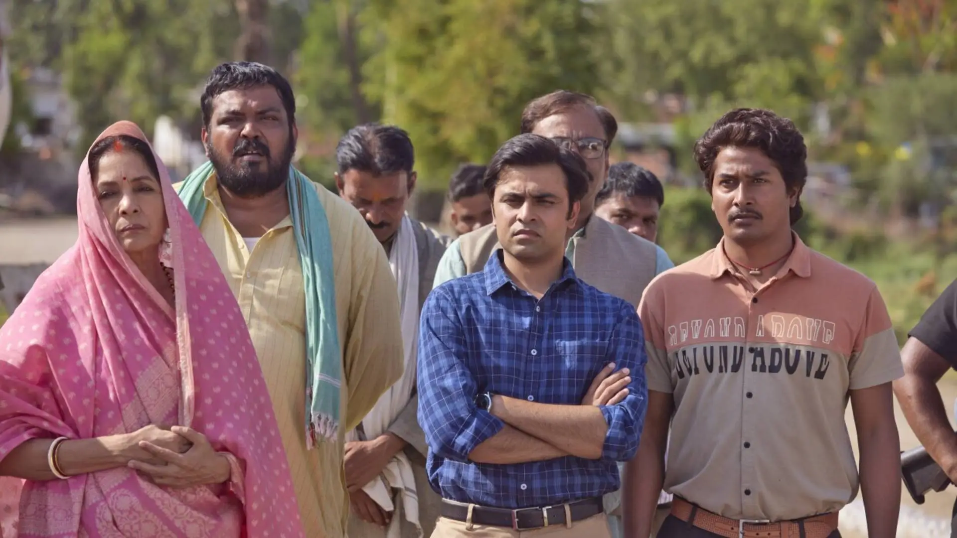 The image is a screenshot of a scene from the tv show, Panchayat. It shows a group of people standing and looking at something in the distance. Among them is the woman who is the village sarpanch. Standing next to her is the deputy pradhan, the panchayat secretary, and his assistant. There are other people standing behind them._Nonprofit humour
