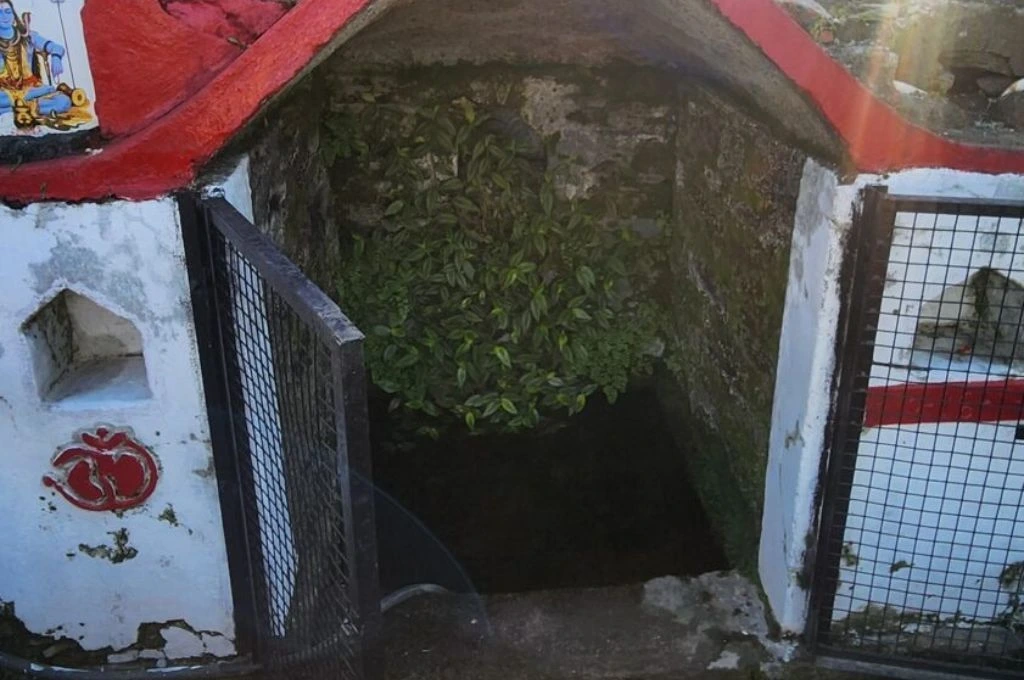The image is a photograph of a naula, or stone-lined reservoir that stores groundwater. The level of the water is lower than the road level. There are plants growing on the walls of the reservoir and it is enclosed by a concrete structure with a gate made of metal net. The walls of the enclosure feature religious motifs._Groundwater conservation