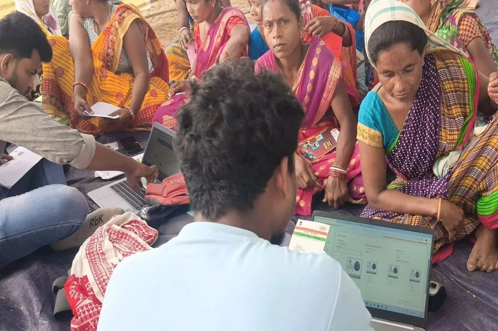 A group of women in colorful sarees sit on the ground as two men use laptops to complete their e-KYC verification for ration cards._Public Distribution System PDS