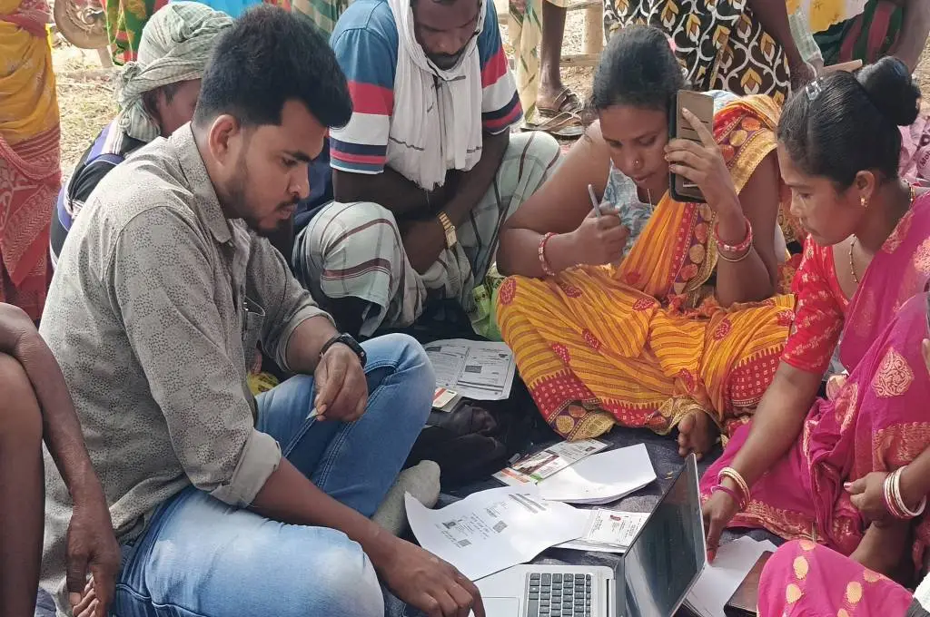 A man and several women sit on the ground, reviewing documents and using a laptop to complete e-KYC verification for ration cards._Public Distribution System PDS