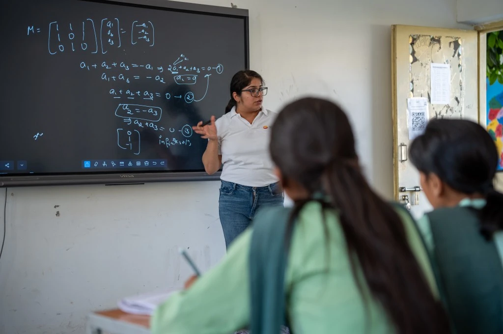 a teacher teaching maths to a class, with mathematical equations on the blackboard behind her and the backs of two girls' heads visible--girls in STEM