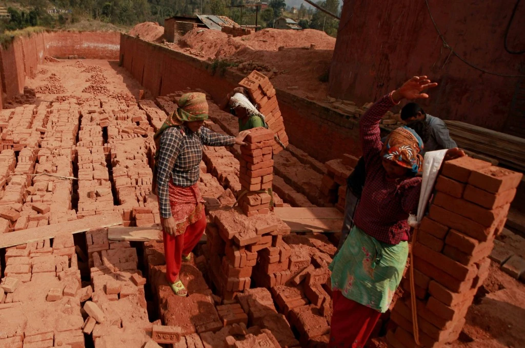 Women carrying stacks of bricks on their backs in a brick kiln, walking carefully over uneven stacks of bricks, with dusty red surroundings and a partly sunlit setting_women workforce participation