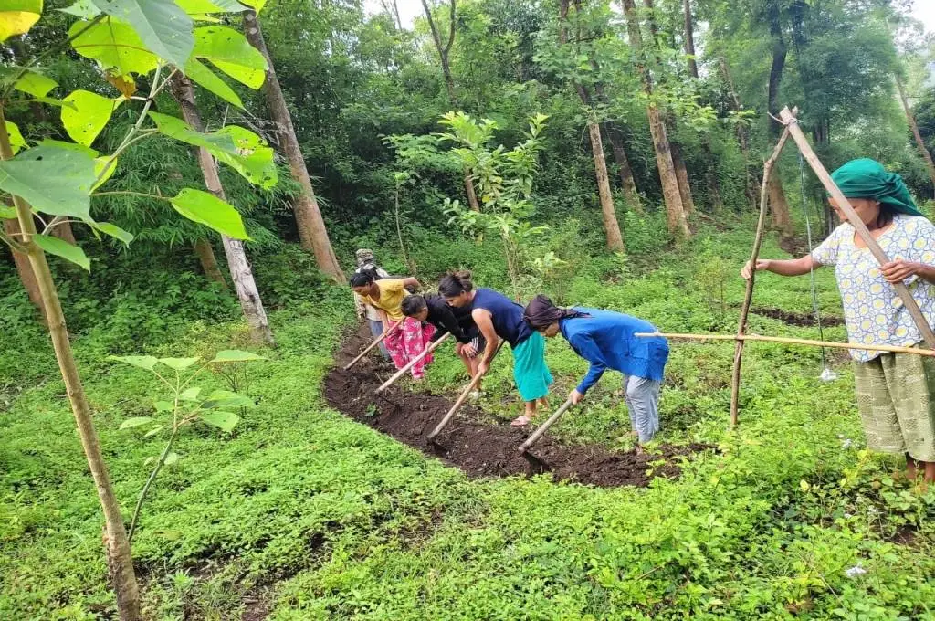 The image shows a group of four women standing in a line in a grass-covered clearing in a forest. Using a shovel, they are digging the soil to create a shallow, horizontal trench. Another woman is standing next to them and she has built a triangular structure using wooden sticks._Kuki-Zo community