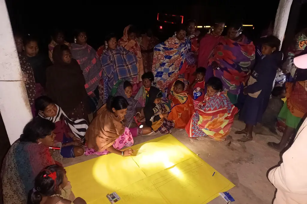 A group of villagers, including women and children wrapped in shawls, gather at night around a large yellow chart paper on the ground, participating in a community mapping exercise under torchlight._sustainable livelihoods