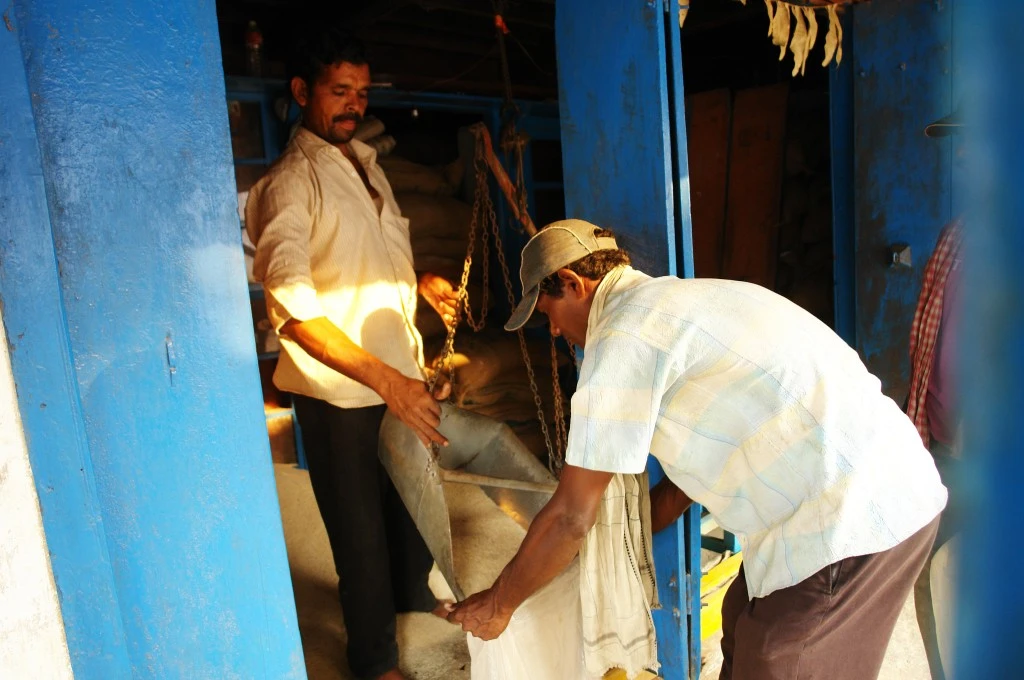 Two men inside a blue-painted shop measure and pour grain into a sack using a metal scoop, at a public distribution system ration shop._Public Distribution System PDS