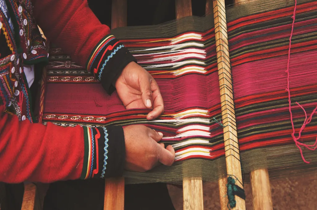 A close-up image of a person weaving colorful fabric on a traditional wooden loom. The individual, dressed in a red garment with embroidered patterns, is using their hands to interlace threads of red, pink, green, white, yellow, and black, creating a detailed textile design._nonprofit acquisitions
