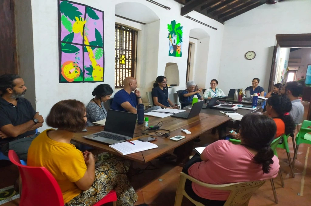 The image features a group of people sitting around two rectangular tables. The people are of different ages and genders, some have laptops and notebooks placed in front of them, and the group is engaged in a discussion. The people are smiling and laughing. The walls of the room have colourful drawings of animals, including a lion and a giraffe, painted on them._Neurodiversity