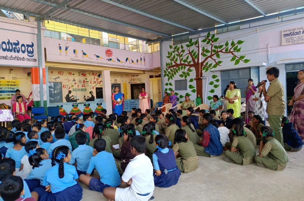 A group of schoolchildren sit on the floor in rows, wearing uniforms in blue and green, facing a stage area where teachers and staff stand. One student holds a microphone and speaks while others listen. The background has colorful educational wall paintings, including science formulas, yoga poses, and a large tree mural with green leaves. Several teachers sit on chairs along the wall, watching the event. The setting appears to be a school assembly or interactive session under a covered courtyard_makkala grama sabhas