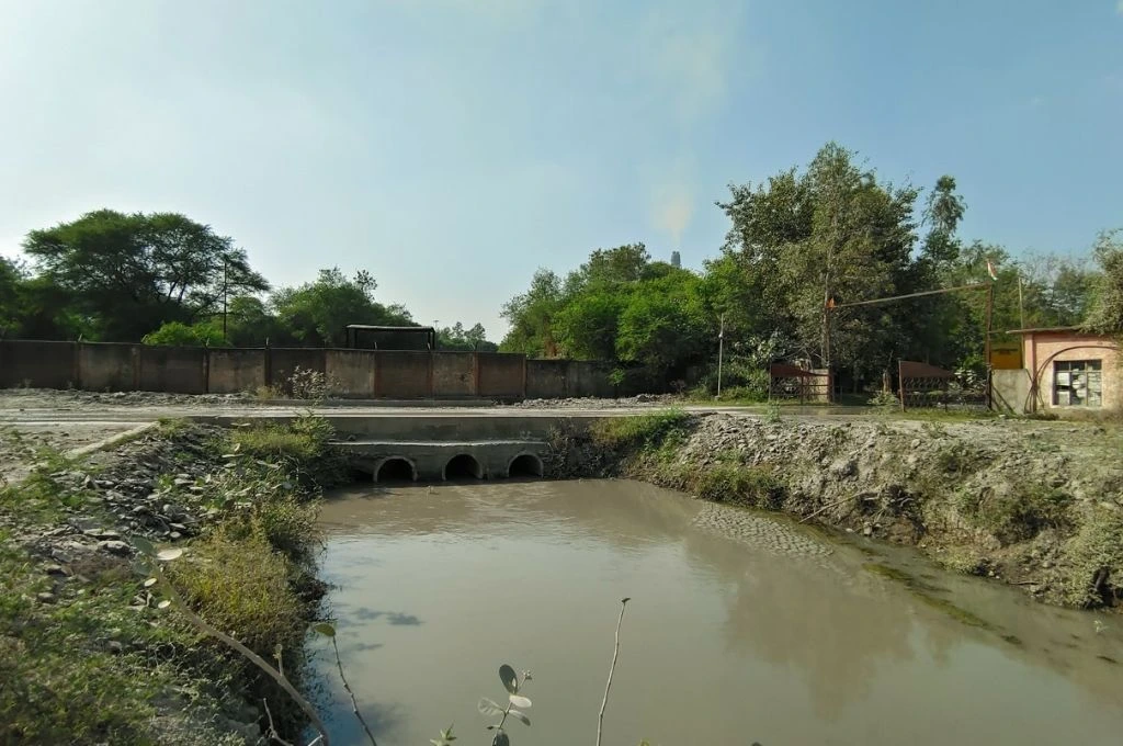 A murky water channel flowing under three concrete culverts, with rocky banks and sparse vegetation in the foreground, and a gated facility surrounded by trees and a high brick wall in the background under a clear sky_enviornment impact assessment