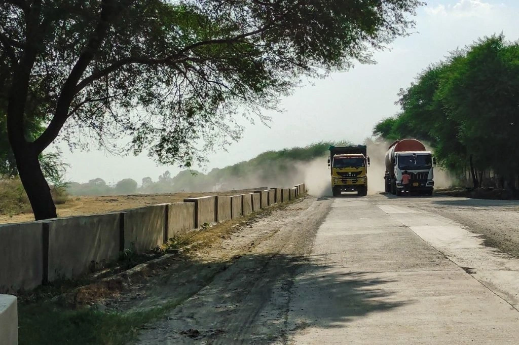 Two heavy trucks drive on a partially paved road, kicking up clouds of dust. Trees line both sides of the road, creating a shaded canopy in parts._ enviornment assessment