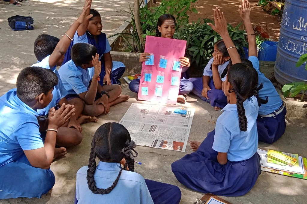 A group of schoolchildren in blue uniforms sit in a circle outdoors on the ground. One girl in the middle holds a pink chart with numbers and pictures, leading the activity. Several children have their hands raised, actively participating in the discussion. Newspapers and notebooks are spread on the ground, indicating a group learning session taking place in an informal setting_makkala gram sabha