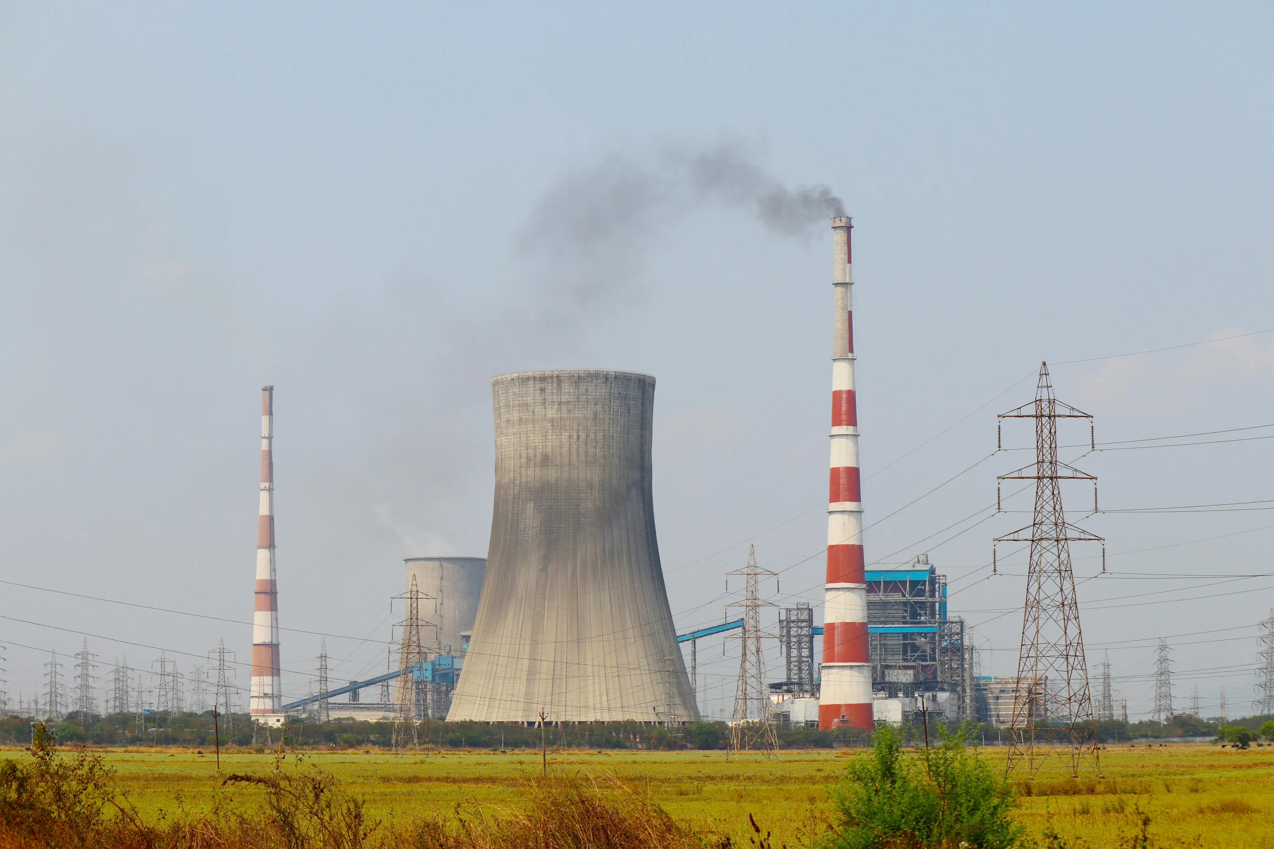 An industrial power plant with tall chimneys emits smoke into the air. Large cooling towers and electricity pylons dominate the scene, surrounded by open fields under a clear sky_enviornment impact assessment