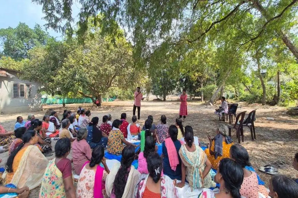 The image is set in a small open ground behind a building. A group of women are sitting on the ground in rows and they are looking ahead at a young man who is standing and addressing the group. Beside him, a young woman is standing. A third woman is seated on a chair to the side._Mental health