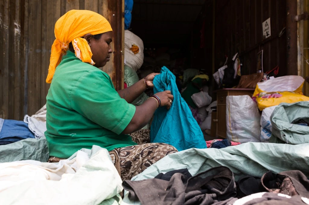 A woman wearing a yellow headscarf and green shirt sits surrounded by piles of clothes, sorting textile waste at a dry waste collection centre._circularity