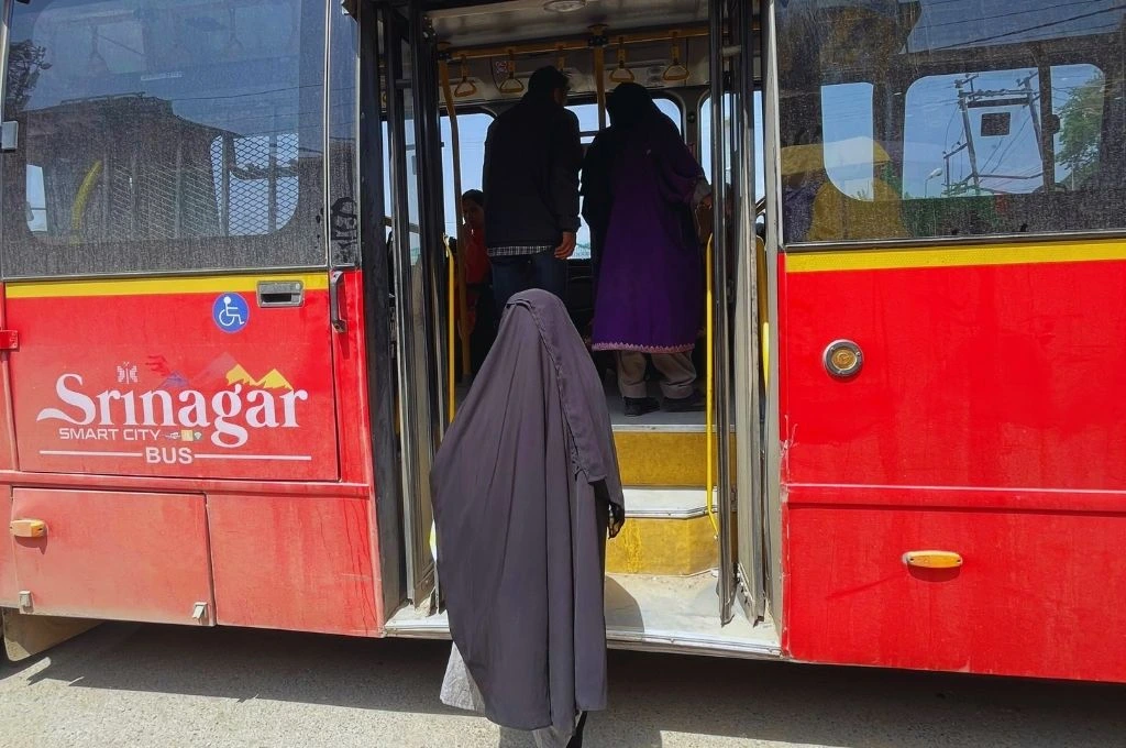 A red public bus labeled “Srinagar Smart City Bus” is parked with its doors open. Several passengers are boarding, including a woman in a full black burqa stepping onto the bus, while others ahead of her are already climbing the steps_women in kashmir