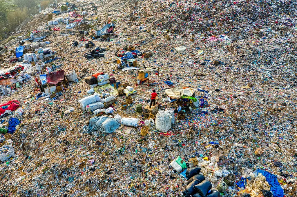 An aerial view of a massive landfill site filled with heaps of waste and plastic debris. Scattered across the trash are makeshift shelters, tarpaulins, and piles of collected recyclables. A few people can be seen sorting through the garbage, surrounded by large sacks and baskets used to gather materials. The scene highlights the harsh working and living conditions of waste pickers amidst an overwhelming expanse of urban waste._Waste management