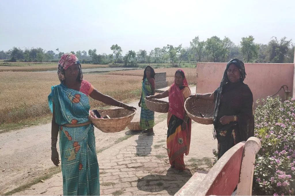 Four women stand outdoors holding empty woven baskets in their hands. They are dressed in colorful sarees and headscarves, standing on a brick path near a pink wall with flowering plants. Behind them, open fields and trees stretch into the background under a clear sky_machines in agriculture