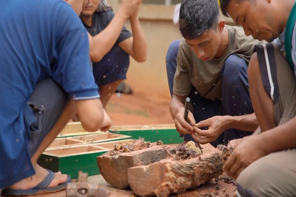 The image shows the author sitting with a group of young boys in a circle on the ground. There are pieces of wooden logs placed on the ground and there are several wooden boxes that have been painted green also placed on the ground. The author is holding a dull iron tool in his hand as he works on something._Beekeeping