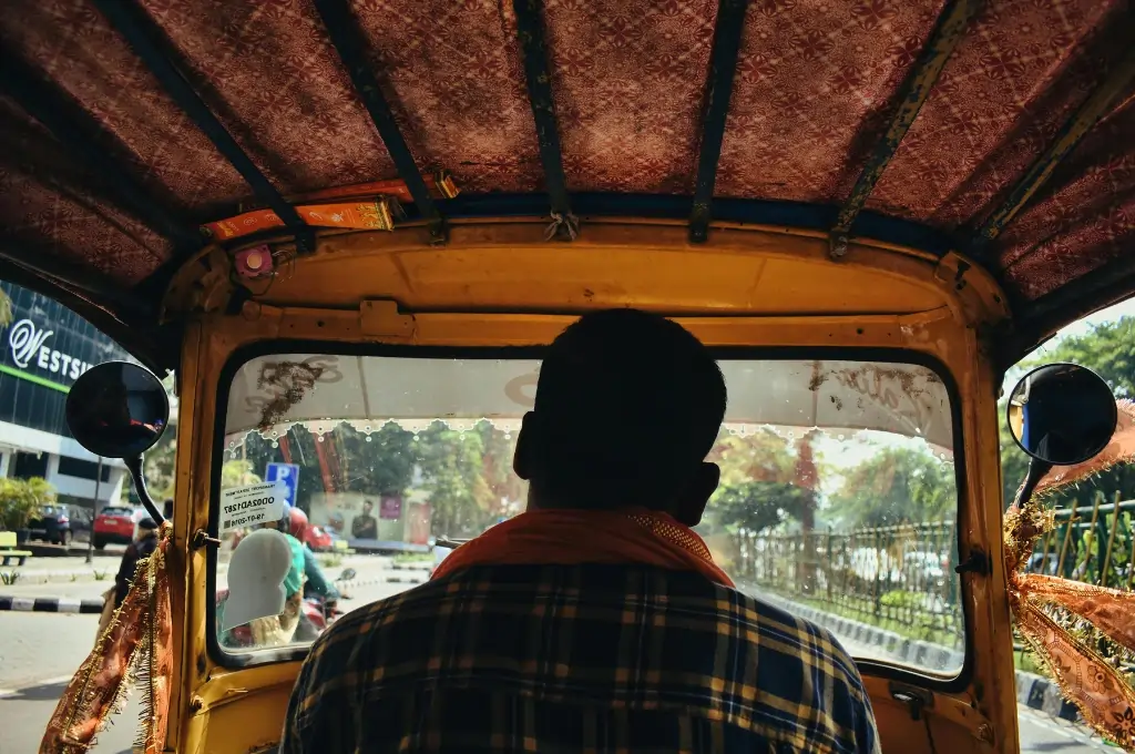 View from inside an auto rickshaw, showing the driver from behind as he drives through a city street with traffic and greenery outside._gig workers