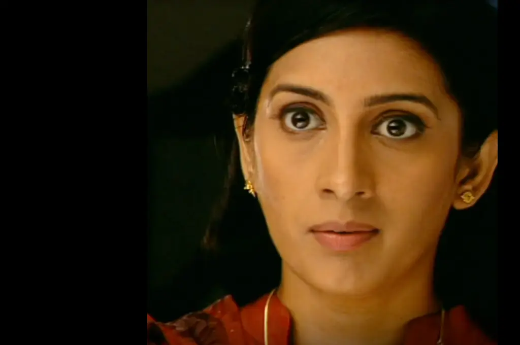 Close-up of Smriti Irani as Tulsi from Kyunki Saas Bhi Kabhi Bahu Thi, with wide, surprised eyes, wearing a red outfit and gold earrings, against a dark background._nonprofit humour