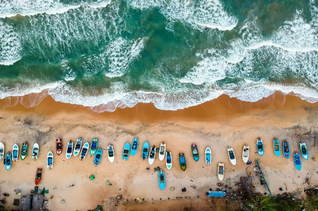 The image is an aerial shot of a beach. The waves from the sea are moving towards the sand, where differently-coloured boats are lined up in a row._Nonprofits