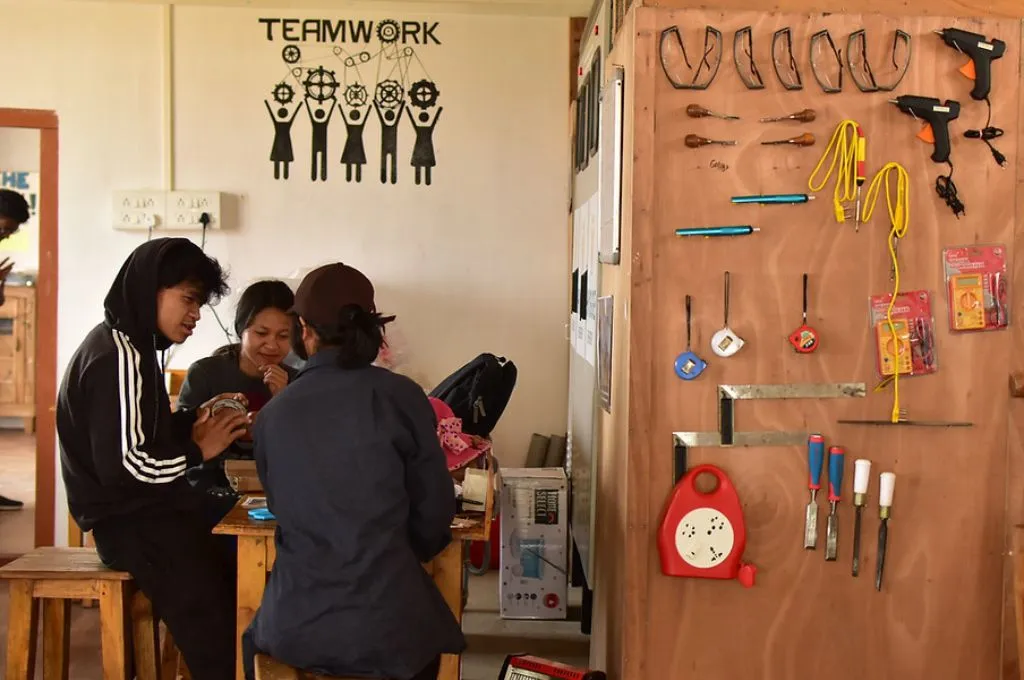 Three young people sitting at a table and talking to each other. They are all wearing black. We can see that the boy is wearing a hoodie with white stripes. To their right on a wooden wall with tools such as measuring tape, drilling machines, screwdrivers etc stick to the wall_government collaboration