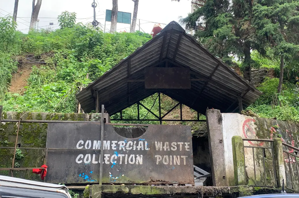 A roofed structure with a weathered sign reading “COMMERCIAL WASTE COLLECTION POINT” stands against a backdrop of greenery and trees._waste management