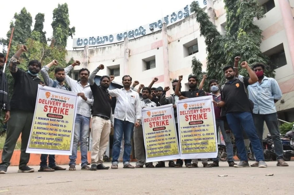 The image shows the author standing with a group of workers. They have their arms and fists raised in a show of solidarity. Some of the workers are carrying posters with the heading, "Swiggy Hyderabad. Delivery Boys Strike.". This is followed by a list of demands including a minimum base pay of INR 35, delivery charge of INR 5, distance pay of INR 12/KM, the removal of 'super zones', and the removal of third parties._Gig workers