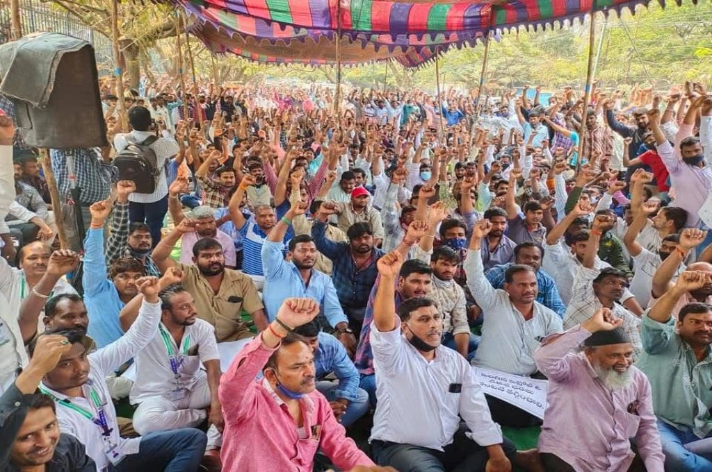 The image shows a huge crowd of people gathered under a tent in an open ground. Everyone has raised their arms up in the air with a closed fist in a sign of solidarity._Gig workers