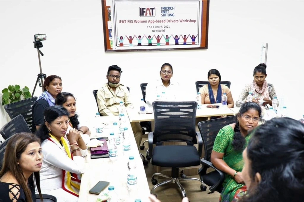 The image shows a group of people sitting around a table and holding a meeting. The wall in the background has bulletin board which has a poster on it. The text on the poster reads, "IFAT-FES Women App-based Drivers Workshop, 12-13 March, 2021. New Delhi"._Gig workers