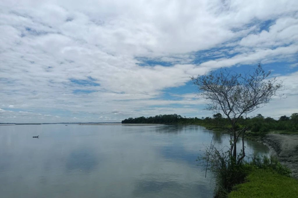 A wide view of a calm river with reflections of the sky, a lone tree on the riverbank, and a mix of blue sky and scattered clouds_Assam herders
