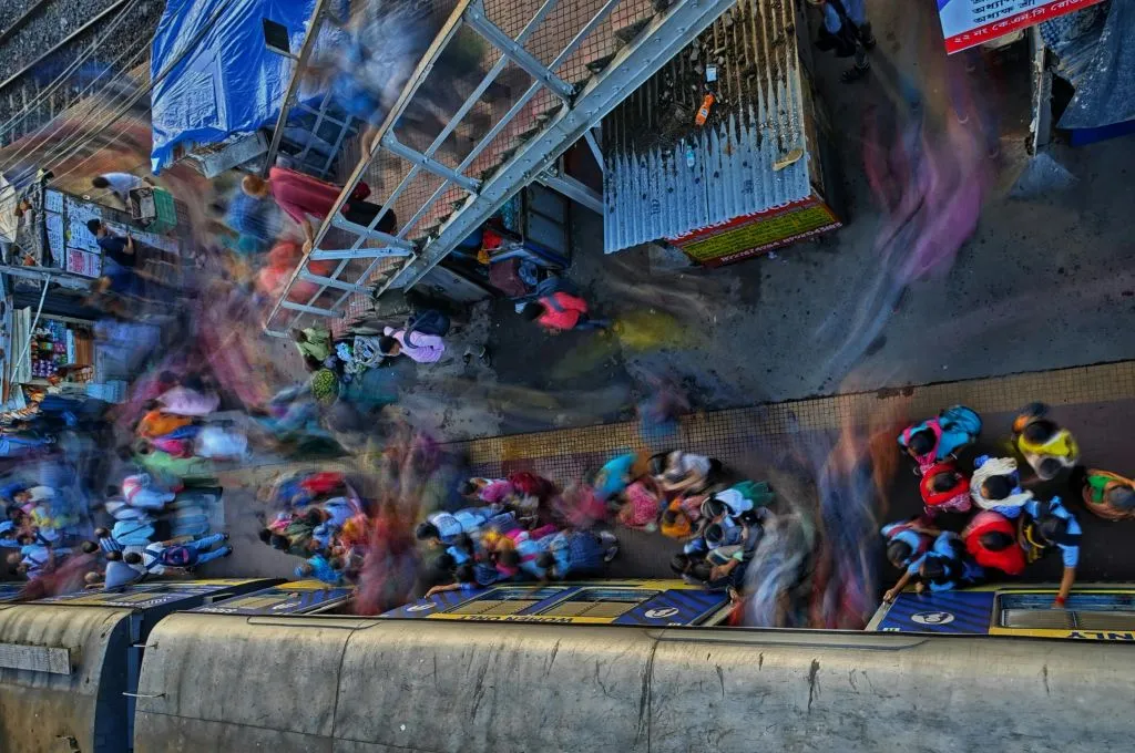The image shows a crowd of people at a railway station platform. A train has stopped at the platform and people are getting on the train while others are de-boarding. Some of the movements are blurred. There are several small shops on the platform that are closed and people are going up and down the staircase in the background._Inequality